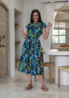 A woman modelling a cap-sleeve cotton midi dress with a vibrant floral and peacock print in teal, green, blue, yellow, and pink on a dark navy background, set in a bright, modern rustic kitchen.