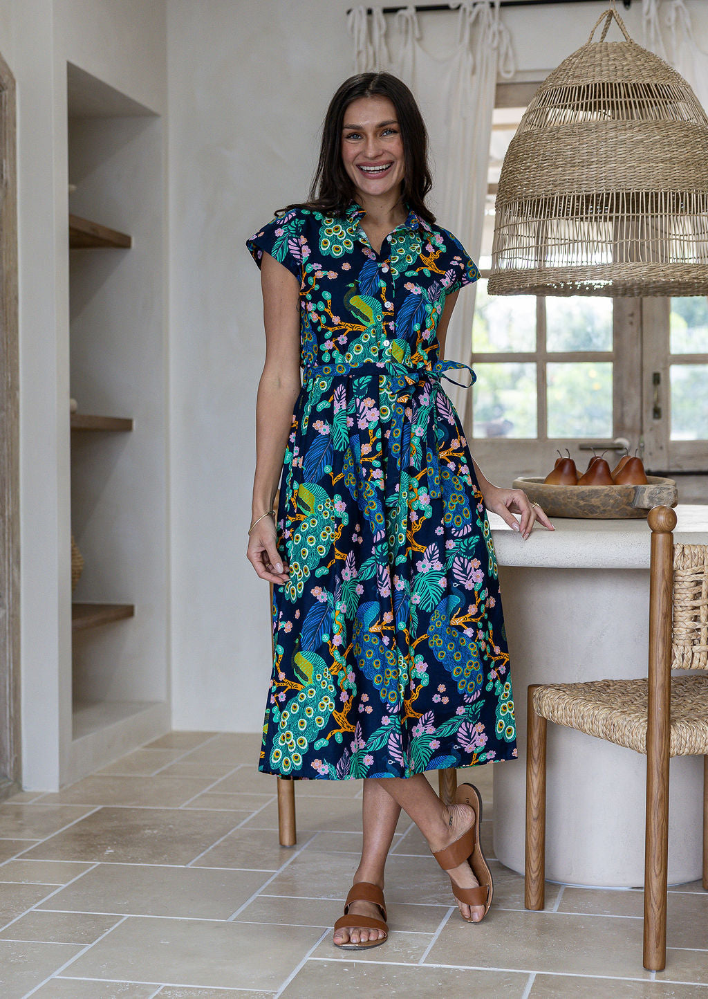 A woman modelling a cap-sleeve cotton midi dress with a collar, buttoned bodice and waist tie, in a vibrant multicolour floral and peacock print