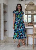A woman modelling a cap-sleeve cotton midi dress with a collar, buttoned bodice and waist tie, in a vibrant multicolour floral and peacock print