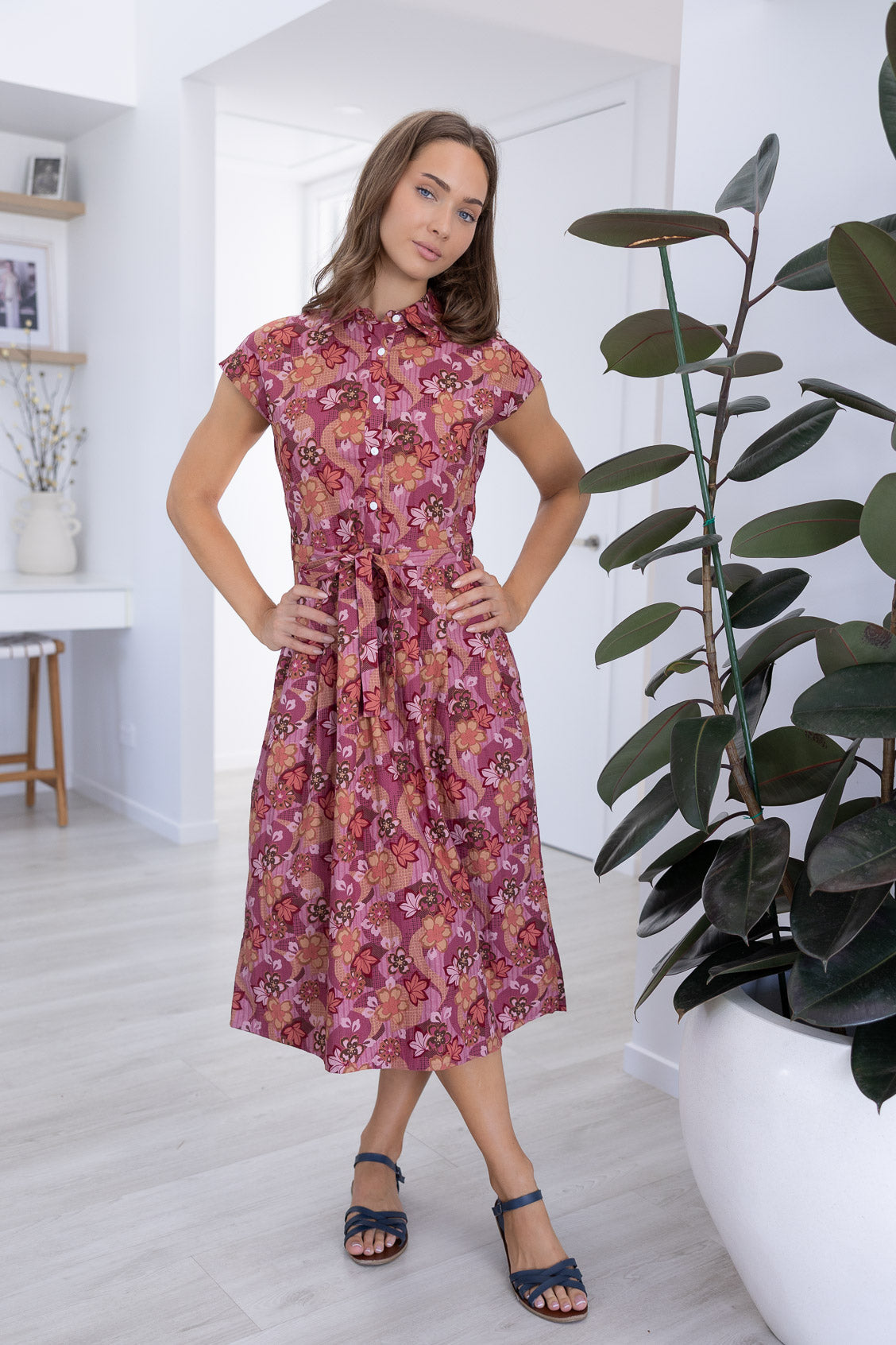 Model stands wearing a midi length floral printed shirt style cotton dress with cap sleeves, buttoned bodice and a removable waist tie