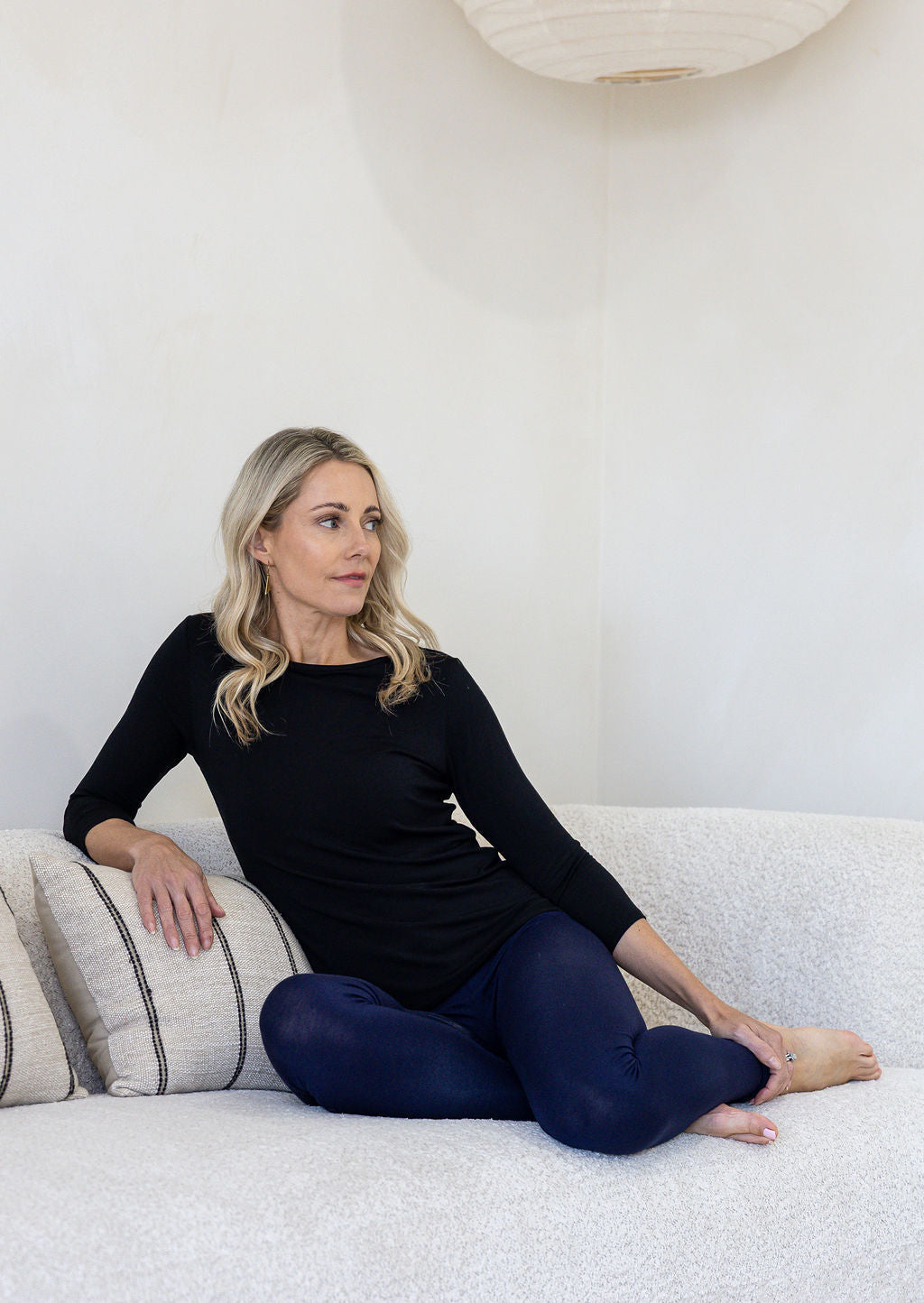 A woman sits on a textured white sofa wearing a black long-sleeve top and dark navy leggings. 