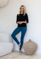Woman wearing high waisted full length soft rayon leggings in teal blue standing in a minimalistic room