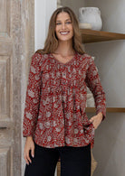 Woman poses in button through cotton shirt in an earthy red based floral print, with her hand in pocket of black pants