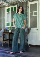 Woman wearing a sage green based floral 100% cotton top and plain pants standing in a rustic indoor setting.