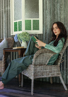 Woman sitting in a wicker chair reading a book in a room with textured walls and a window.