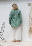 Woman stands backwards wearing a cotton long sleeve shirt that is longer at the back, paired with plain cotton pants