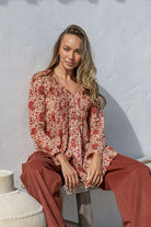 Woman sitting on a stone block wearing a patterned blouse and red pants against a light gray wall.