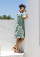 Woman stands sideways wearing a relaxed fit, sage green based floral cotton print dress with pockets and empire waistline. 