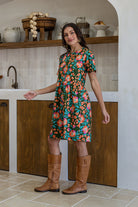 Woman stands sideways wearing a relaxed fit, olive green based floral cotton print dress with pockets and empire waistline. 