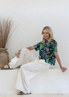 Woman sits on a white ledge in a bright, minimalist room, wearing a short-sleeve 100% cotton top paired with white plain pants