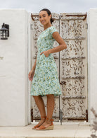 Model with one hand in pocket wears a knee length A line dress with cap sleeves in sage green based floral print
