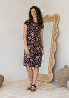 Model standing in a 100% cotton dress in Indian traditional floral print cotton dress with with rust, tan and sand tones on a black base.