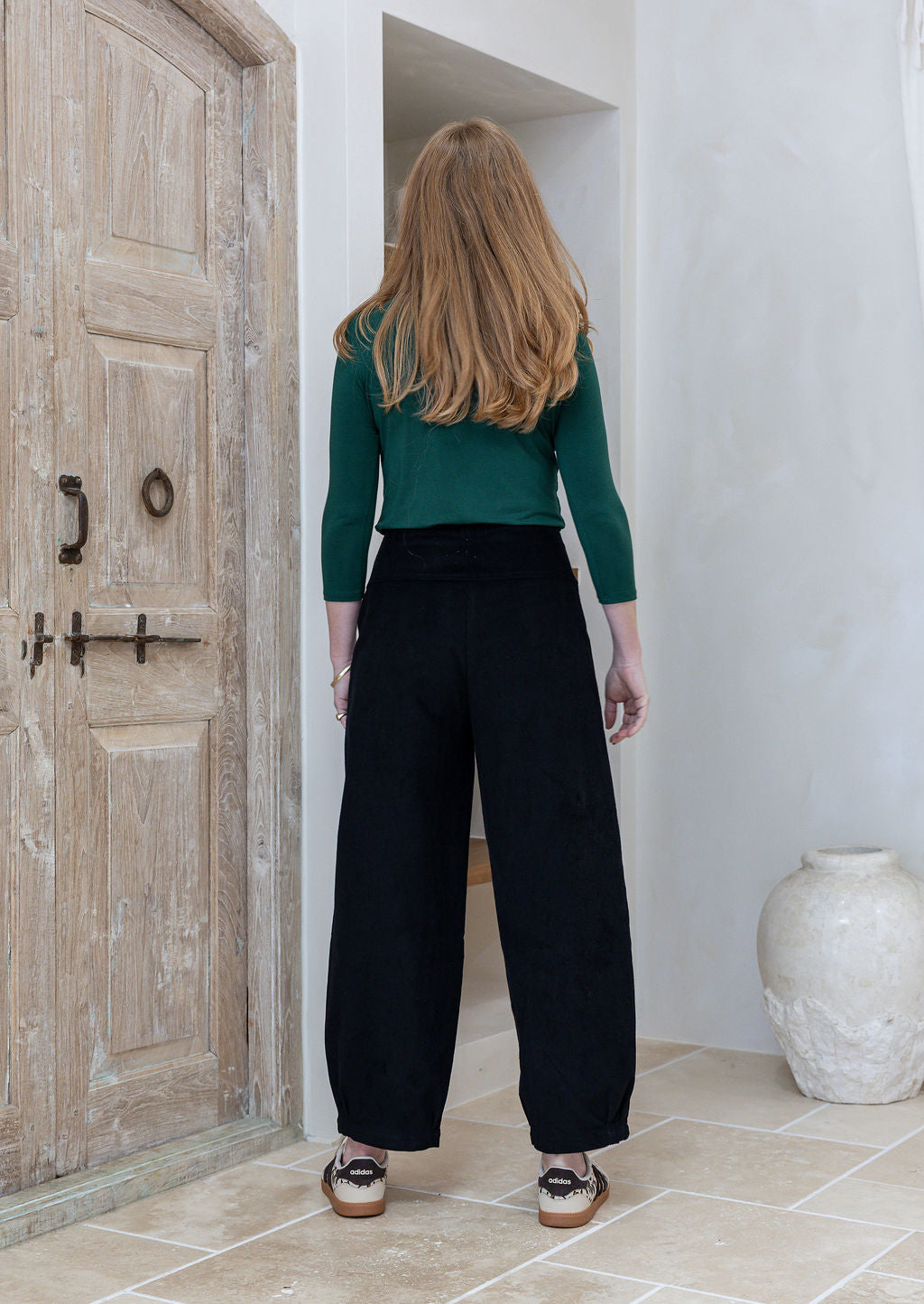Woman stands backwards wearing a black cotton corduroy pants paired with green basic top