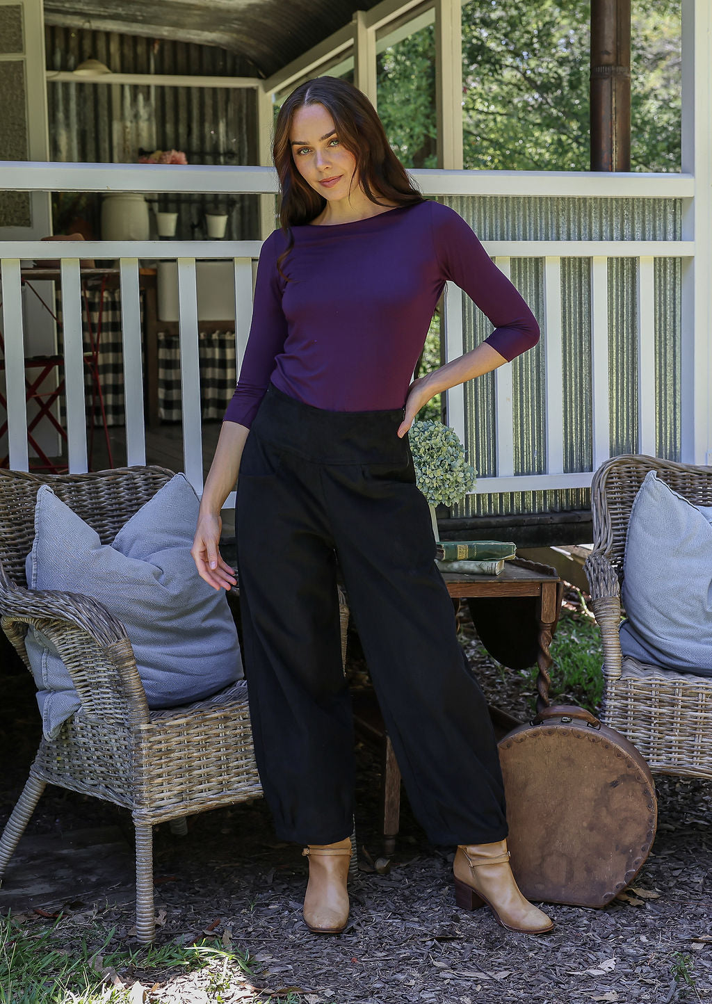 Woman in a purple top and black cotton corduroy pants standing in front of a porc and wicker chairs