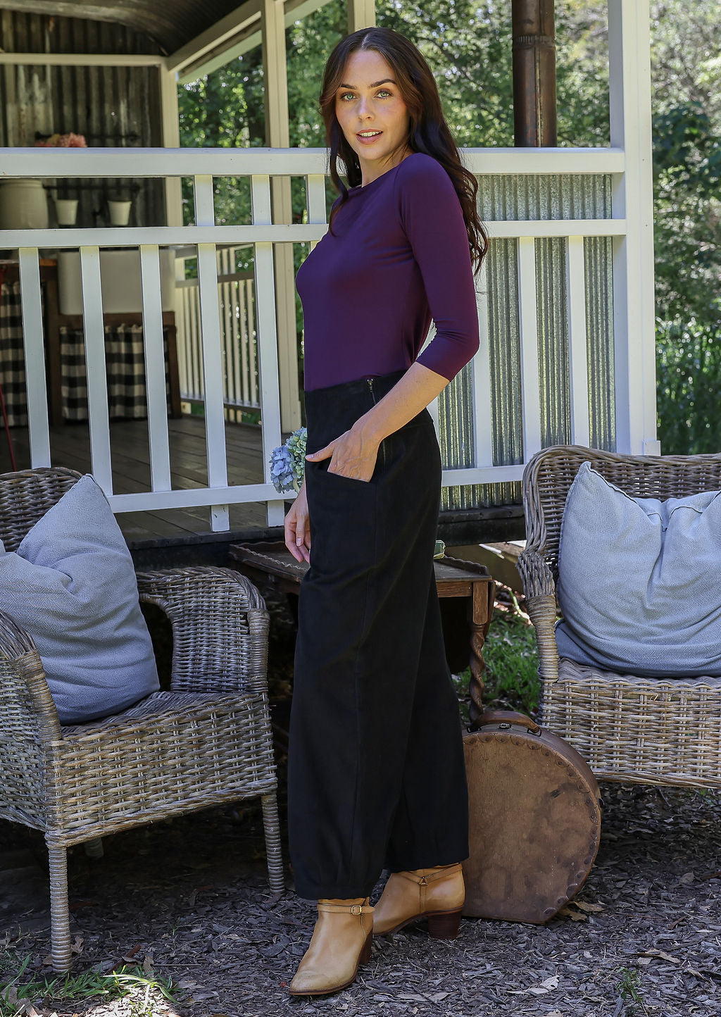 Woman stands side on in a purple top and black cotton corduroy pants with a side zip with hand in pocket