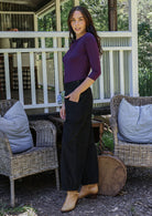 Woman stands side on in a purple top and black cotton corduroy pants with a side zip with hand in pocket