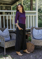 Woman looks over shoulder wearing a purple stretch rayon top and black cotton corduroy pants with hands in pockets