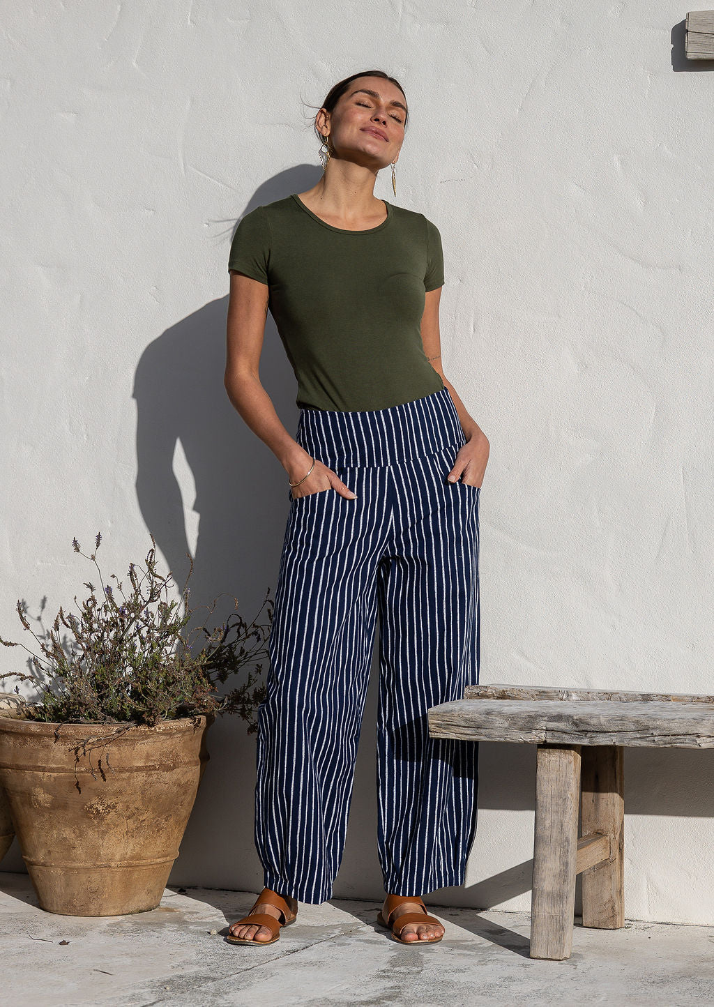 Woman posing in 100% cotton wide-leg navy based white strioed printed pants with wide flat waistband, side zipper and small pleats at the ankles.