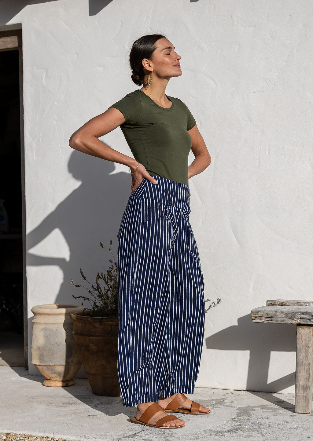 Model standing sideways wears 100% cotton wide leg pants with side zipper, pockets and small pleats at ankles.