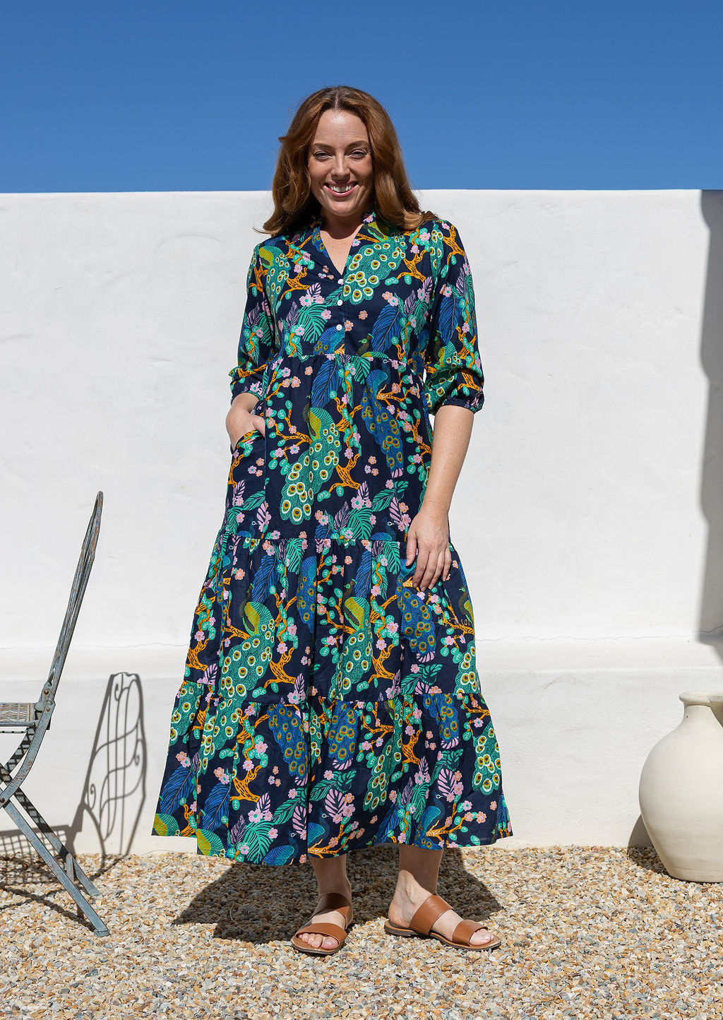 A woman wearing a 3/4 sleeved, buttoned bodice cotton maxi dress with a multicolour floral and peacock print, with hand in pocket