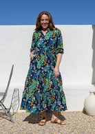 A woman wearing a 3/4 sleeved, buttoned bodice cotton maxi dress with a multicolour floral and peacock print, with hand in pocket