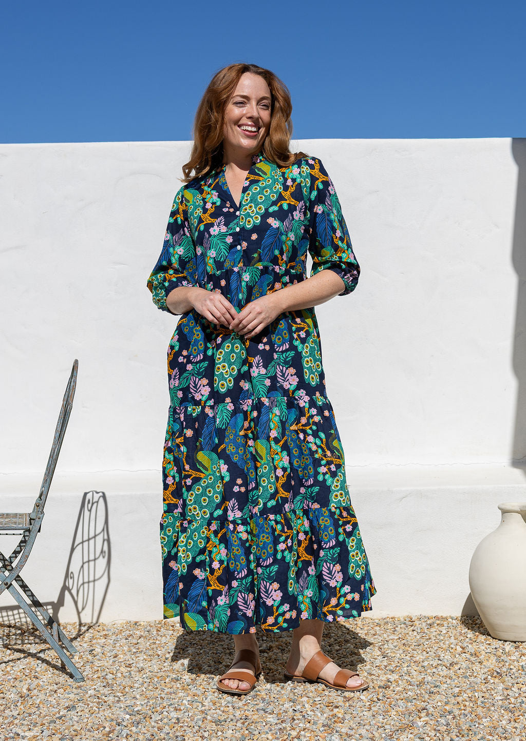 A woman wearing a 3/4 sleeved maxi dress with a vibrant multicolour floral and peacock print on a dark navy background, standing outdoors against a white wall under a clear blue sky.
