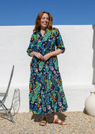 A woman wearing a 3/4 sleeved maxi dress with a vibrant multicolour floral and peacock print on a dark navy background, standing outdoors against a white wall under a clear blue sky.