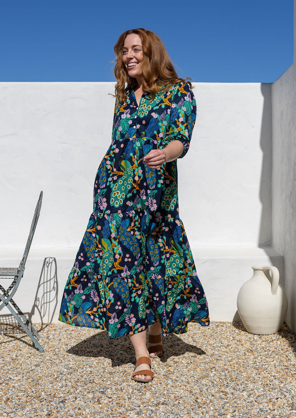A woman wearing a 3/4 sleeved maxi dress with a floral and peacock print in shades of teal, green, blue, orange and pink, walking outdoors against a white wall.