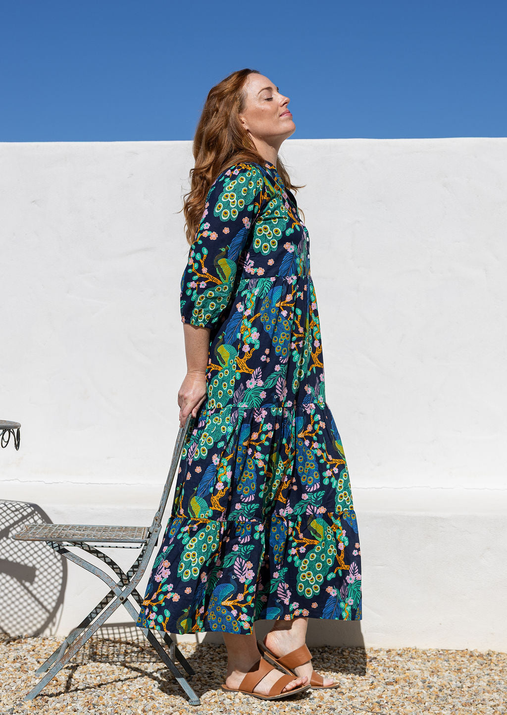 A woman leaning on a chair wearing a 3/4 sleeved cotton maxi dress with a colourful floral and peacock print in teal, green, yellow, and pink.