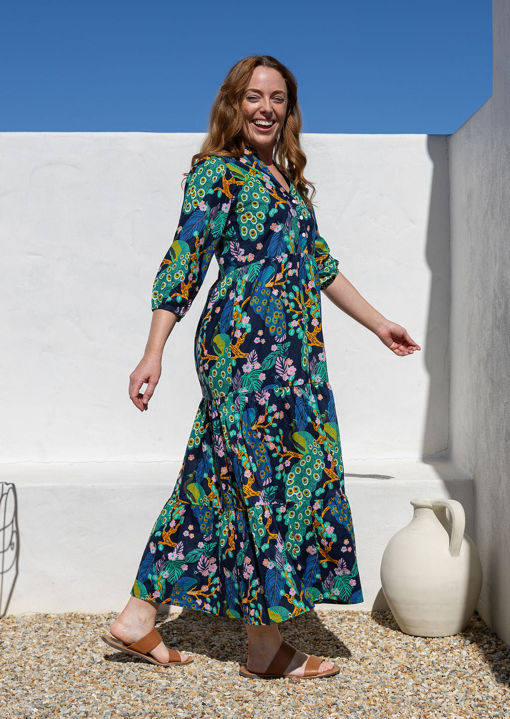 Woman standing side on wearing a 3/4 sleeve cotton maxi dress with a multicoloured peacock print, standing outdoors against a white wall and blue sky.