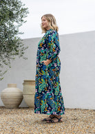 A woman stands sideways wearing a 3/4 sleeved cotton maxi dress with a colourful floral and peacock print in teal, green, yellow, and pink.