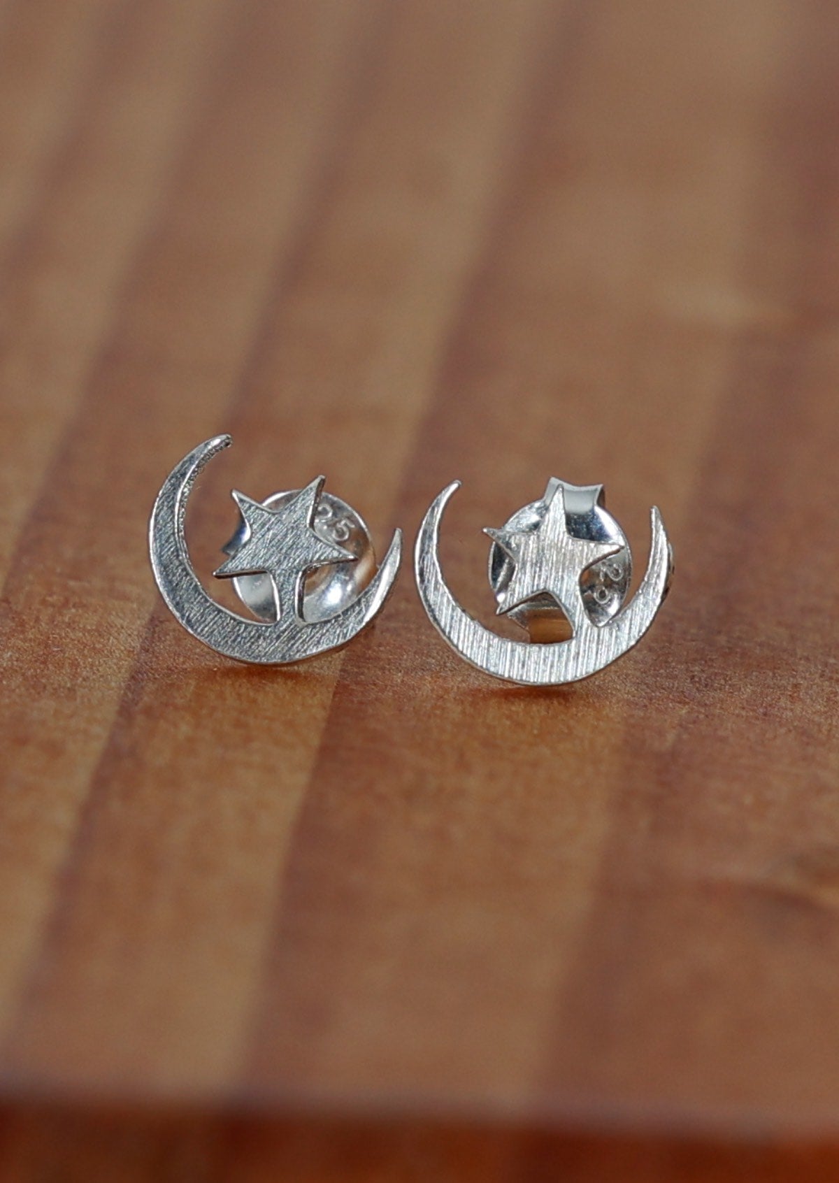 A close-up image Moon and Stars silver stud earrings shaped like a crescent moon with a star inside it on a wooden table.