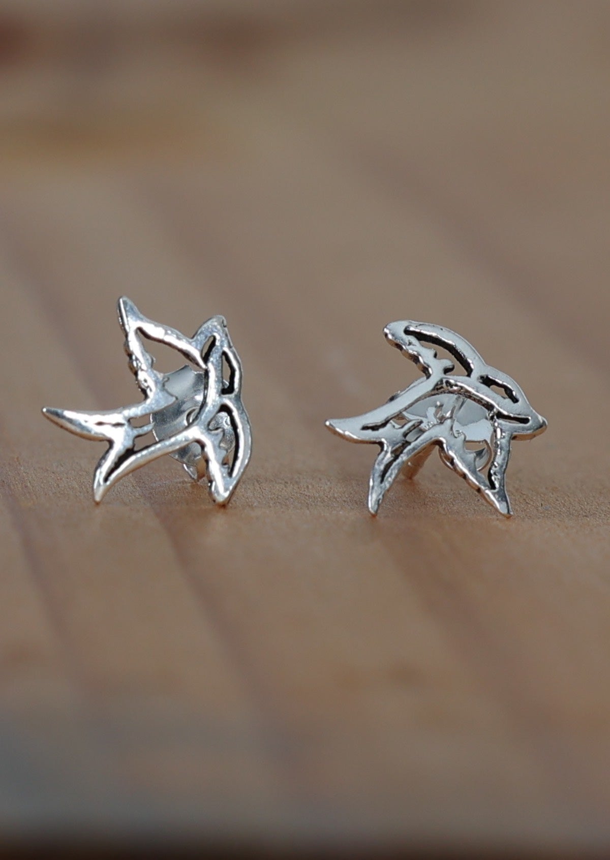 A close-up image of silver Swallow stud earrings in the shape of a swallow bird on a wooden surface.