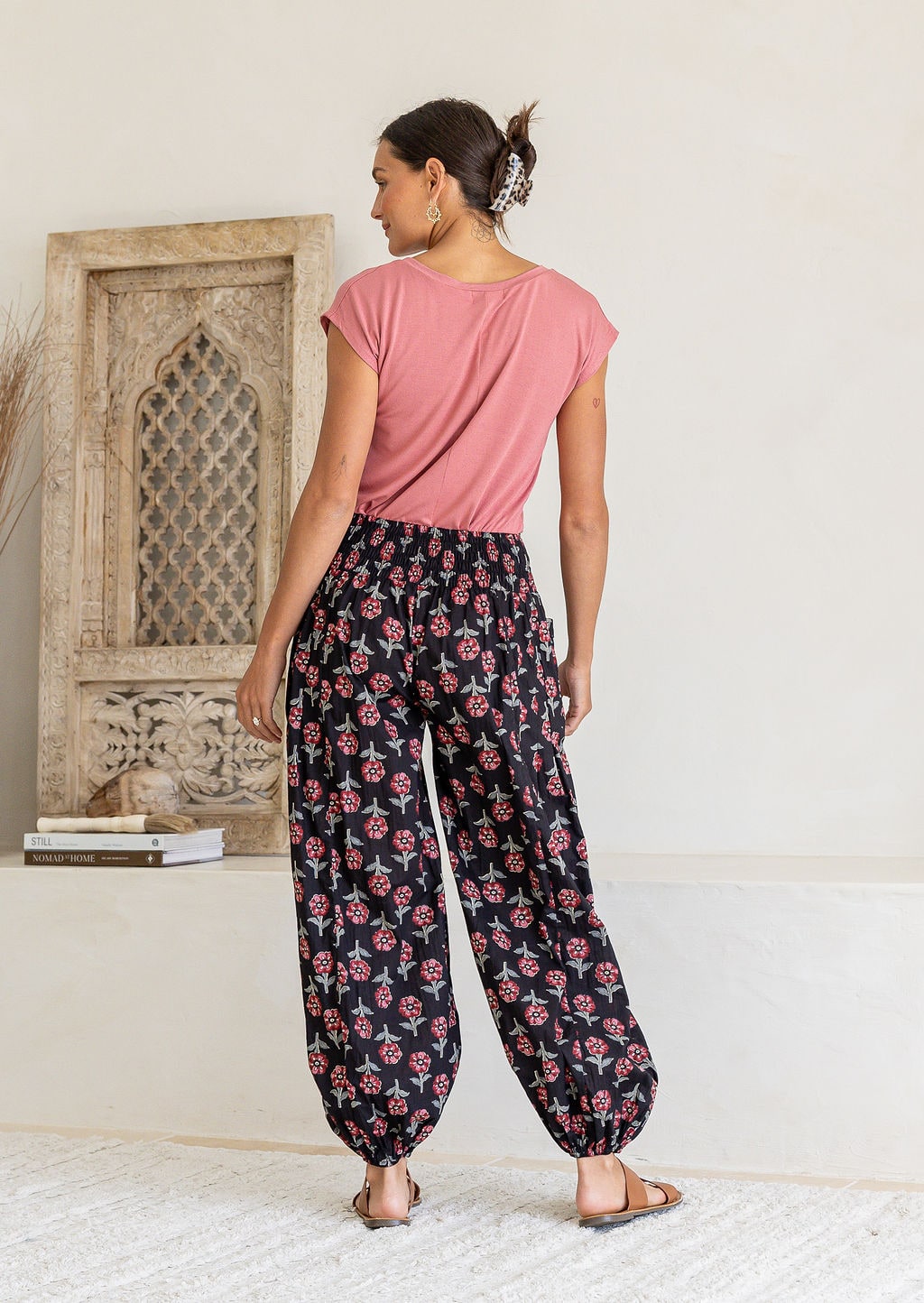 Woman stands backwards wearing a basic top and blue based floral pants with wide elastic waist and ankles.