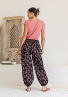 Woman stands backwards wearing a basic top and blue based floral pants with wide elastic waist and ankles.