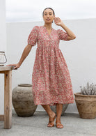 Woman wearing a pink floral cotton dress standing in an outdoor setting with plants and a table.