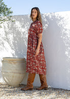 Woman standing side on in a red floral cotton midi length dress standing in front of a white wall.