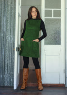Woman wearing a green cotton corduroy sleeveless tunic layered over a black top and leggings and tan boots, with hands in pockets