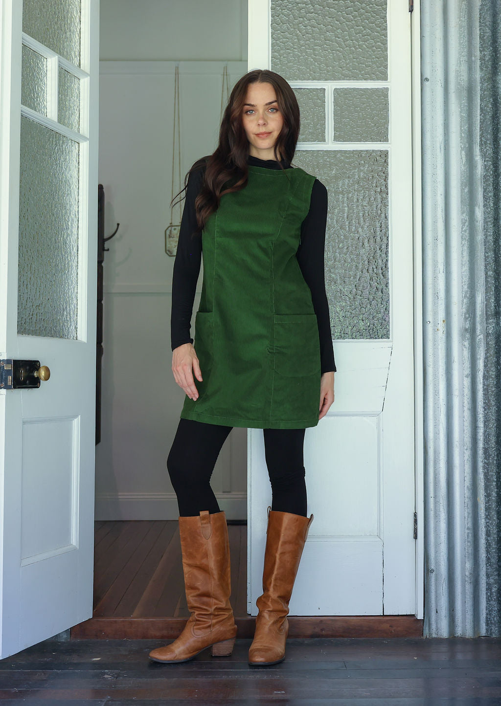 Woman wearing a green cotton corduroy sleeveless tunic layered over a black top and leggings and tan boots