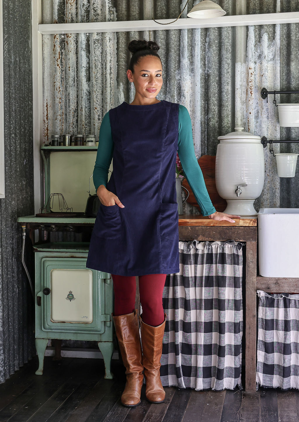 Woman in a navy blue dress standing in a rustic kitchen with checkered curtains and vintage stove.