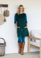 Woman wearing a dark green sleeveless cotton tunic over a teal long-sleeve top and leggings, and brown knee-high boots, with hands in pockets