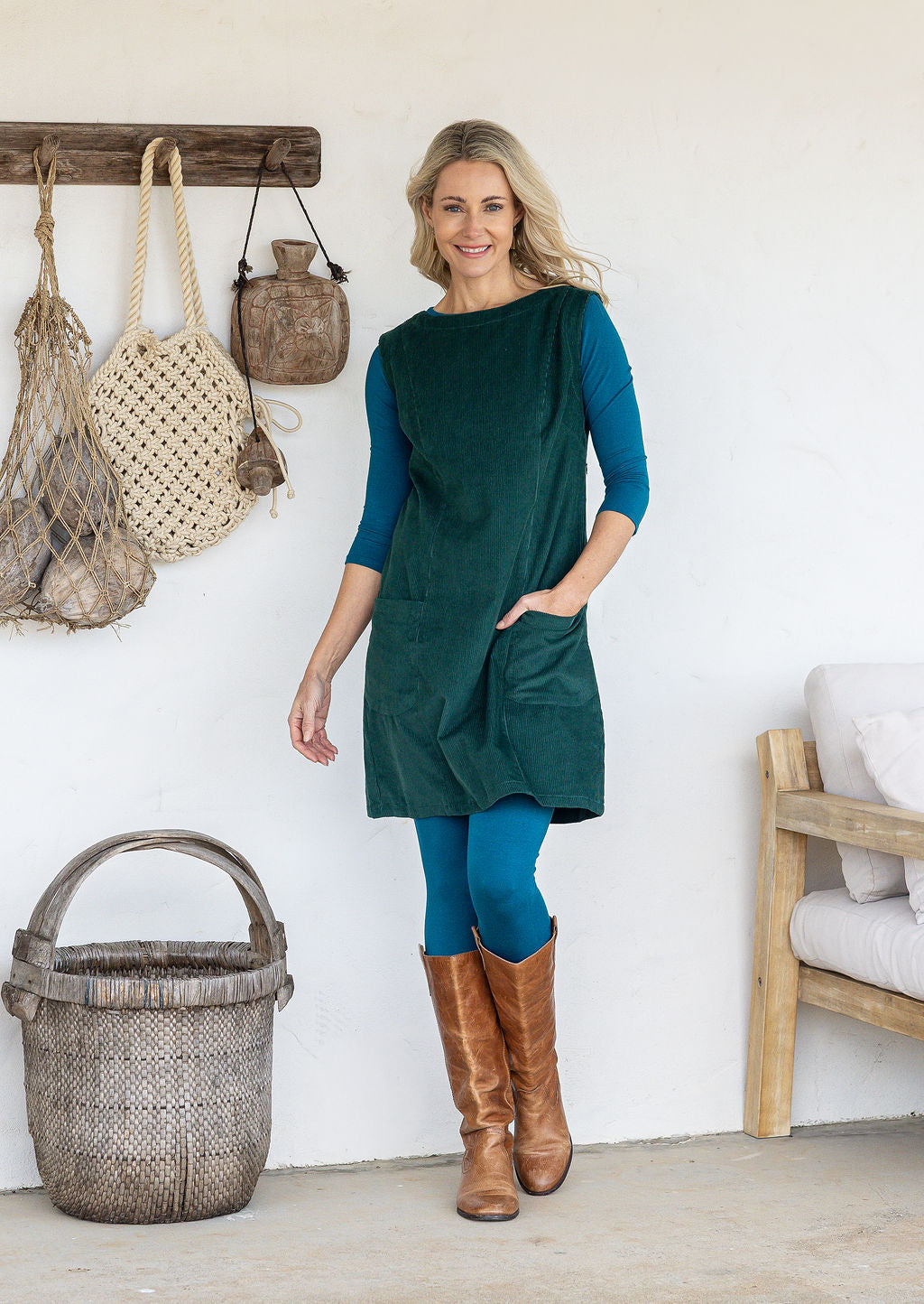 Woman wearing a sleeveless dark green cotton tunic over teal long-sleeve top and leggings and tan knee-high boots, with one hand in pocket