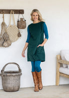 Woman wearing a sleeveless dark green cotton tunic over teal long-sleeve top and leggings and tan knee-high boots, with one hand in pocket