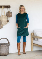 A woman wearing a dark green sleeveless cotton pinafore dress over a teal long-sleeve top and leggings, paired with brown knee-high boots, with hands in pockets