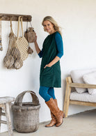 A woman wearing a dark green cotton corduroy tunic over a teal long-sleeve top and leggings, standing in a rustic room with a woven basket, wooden furniture