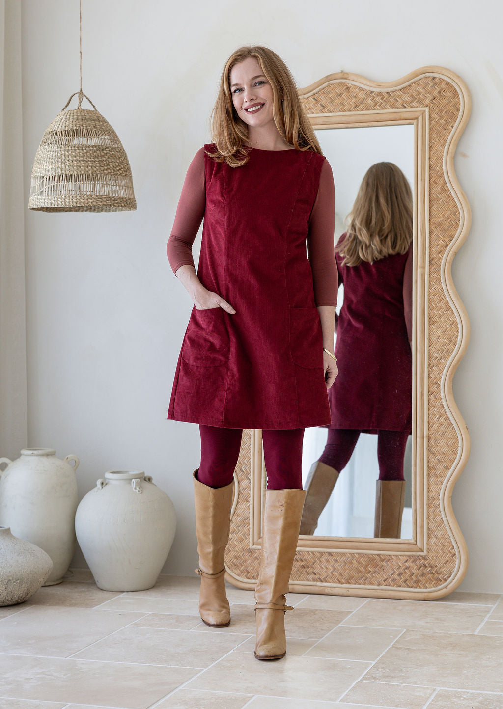 Woman poses in cotton corduroy tunic in cabernet with one hand in pockets styled with leggings and tan boots