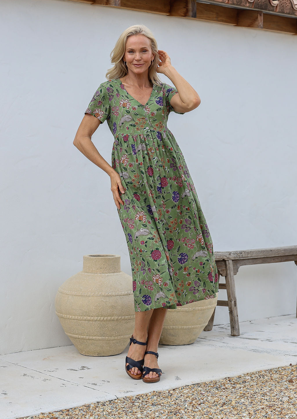 Woman wearing relaxed fit button through dress in moss green based multicoloured floral print with V neckline, pockets and short sleeves.