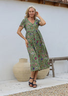 Woman wearing relaxed fit button through dress in moss green based multicoloured floral print with V neckline, pockets and short sleeves.