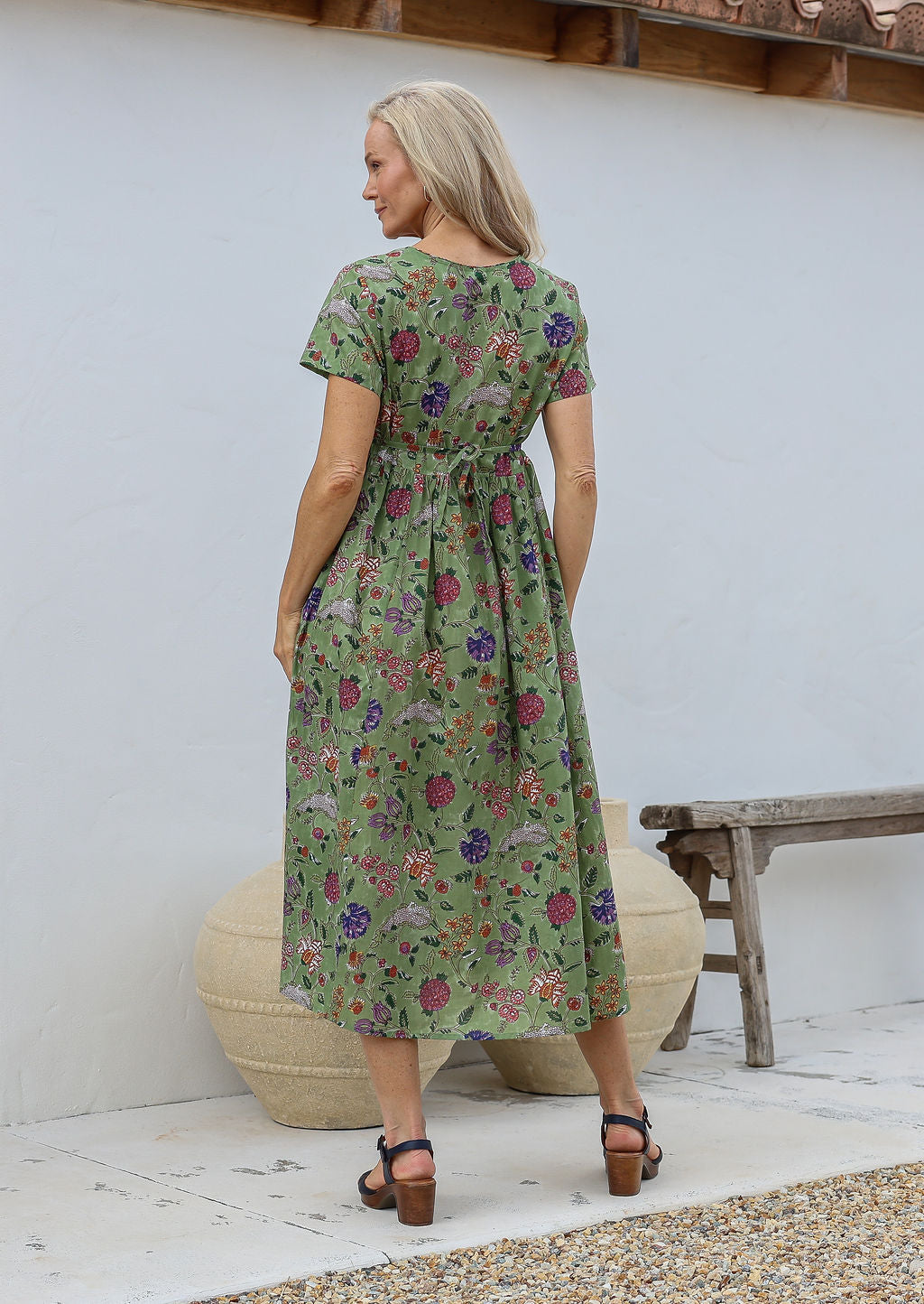 Woman standing in a back pose wearing a 100% cotton relaxed fit dress in moss green with short sleeves, pockets and drawstring waist tie.
