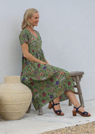 Woman sitting on a wooden bench wearing a lightweight cotton moss green based floral print dress with short sleeves, v-neckline and pockets.